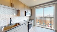 This contemporary single-floor unit offers a thoughtfully designed layout with modern finishes throughout. The kitchen serves as a central highlight, featuring sage-toned shaker-style cabinetry, dark countertops, and a striking white herringbone tile backsplash. Stainless steel appliances — including a refrigerator, dishwasher, over-the-range microwave, and electric range — complete the well-equipped cooking space. Light wood-look flooring flows seamlessly throughout the unit, creating a cohesive and airy aesthetic.

A large sliding glass door in the kitchen and living area opens to a private balcony, flooding the interior with natural light and offering open views of the surrounding cityscape. A second window in the living space further enhances the brightness of the room.

The hallway connects the main living areas to the private spaces and features a stylish sliding barn door with matte black hardware, adding a contemporary farmhouse touch. The bathroom includes a full soaking tub and shower combo, finished with floor-to-ceiling vertical white subway tile and matte black fixtures, lending a clean and classic look. Recessed lighting throughout the unit ensures consistent illumination, while the neutral color palette and modern hardware choices create a polished and cohesive living environment.