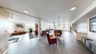 The 7th Floor Sky Lounge at 18004 Midvale Ave in Shoreline, Washington, is a thoughtfully designed communal amenity space offering an elevated retreat with sweeping views of the surrounding area. The open-plan layout seamlessly connects multiple distinct seating and gathering zones, all unified by a striking herringbone-patterned flooring that runs throughout.

The main lounge area is anchored by a sleek linear fireplace framed beneath a wall-mounted television, surrounded by plush seating including deep sofas, tufted armchairs in rich amber and burgundy tones, and velvet accent chairs in bold orange. A cozy nook features built-in banquette seating with colorful throw pillows, tufted ottomans, and small café-style tables — ideal for casual conversation or quiet relaxation. Large windows on multiple sides of the space flood the interior with natural light and provide views of the Shoreline skyline and surrounding greenery.

The full catering kitchen is a standout feature, complete with stone countertops, light wood cabinetry, stainless steel appliances, a built-in dishwasher, and an expansive island topped with pendant globe lighting — well-suited for events and gatherings. A dining area with a long table and modern chairs sits adjacent to the kitchen, offering a functional space for group dining. Accent walls featuring warm, textured finishes and curated artwork add visual depth, while recessed lighting and interior greenery contribute to an inviting, upscale ambiance throughout.