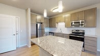 Located at 146 Westgate Mall in Madison, Wisconsin, this single-floor residence offers a thoughtfully designed open-concept layout with contemporary finishes throughout. The kitchen serves as the centerpiece of the living area, showcasing a large granite-topped island, warm-toned cabinetry, a classic subway tile backsplash, and a full suite of stainless steel appliances including a refrigerator, range, microwave, and dishwasher. Pendant lighting and a flush-mount fixture illuminate the space, while light hardwood flooring flows seamlessly throughout the main living areas. The kitchen opens directly to the living area, which is outfitted with a ceiling fan and sliding glass doors leading to a private balcony with exterior views. The bedrooms are carpeted for comfort; one features a large picture window with an urban view and a ceiling fan, while another includes a spacious walk-in closet with built-in shelving. The bathroom is finished with granite countertops, warm wood cabinetry, a wide vanity mirror with bar lighting, and coordinating light hardwood flooring. This residence combines functional design with quality materials to create a clean, modern living environment.