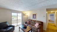 This single-floor unit offers a practical and comfortable layout across several distinct living spaces. The living room serves as the heart of the home, featuring a textured ceiling, carpet flooring, and a sliding glass door that opens to a private balcony with outdoor views — allowing natural light to fill the room. A wall-mounted air conditioning unit ensures year-round comfort.

Adjacent to the living room, the dining area provides a dedicated space for meals, highlighted by a ceiling fan with a light fixture overhead and ample room for a full dining set.

The kitchen is efficiently designed with dark cabinetry, light countertops, tile flooring, a stainless steel sink, a dishwasher, and a white electric range with an overhead exhaust hood — offering all the essentials for meal preparation.

The bedroom is a calm retreat with carpet flooring, a window with blinds for natural light and privacy, and sufficient space for a full furniture arrangement.

A dedicated home office features carpet flooring, a window overlooking greenery, and enough space to accommodate a large L-shaped desk setup — ideal for remote work or study.

The full bathroom is clean and functional, featuring a tiled tub-shower combination, a vanity with storage, and tile flooring throughout.