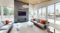 Located at 16017 60th St E in Sumner, Washington, this expansive single-story amenity facility offers a thoughtfully designed, multi-functional layout across a generously sized floor plate. Light-toned hardwood flooring flows continuously throughout, unifying the various activity zones and contributing to a bright, cohesive aesthetic.

The main lounge area serves as an architectural centerpiece, featuring soaring ceilings with a striking wood-paneled soffit, floor-to-ceiling windows that invite abundant natural light and outdoor views, and a full-height tile fireplace wall with an integrated TV. A sculptural pendant light anchors the seating area below.

Adjacent to the lounge is a well-appointed kitchen with dark slate cabinetry, stainless steel appliances, and ample counter space. A dining area with a large wooden table and white spindle chairs sits nearby, illuminated by a trio of modern pendant lights.

A secondary social zone features bold red accent walls with built-in bench seating, a billiards table, and a lounge seating area with a wall-mounted fireplace near the entry. Colorful geometric artwork decorates a dedicated gallery wall. A fitness room with equipment and large windows rounds out the facility, offering a complete, amenity-rich environment.