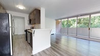 Located at 801 Cooper Landing Rd in Cherry Hill, New Jersey, this single-floor residence offers a thoughtfully designed open-concept layout that seamlessly blends everyday functionality with a clean, modern aesthetic. The kitchen is outfitted with dark espresso cabinetry, a gas range, stainless steel appliances, and a breakfast bar with seating overhang — an ideal setup for casual dining and entertaining. Tile flooring in the kitchen transitions smoothly into the wide-plank hardwood-style flooring of the living area, creating a cohesive visual flow throughout the space. The living area is flooded with natural light courtesy of a floor-to-ceiling sliding glass window wall, which frames lush, tree-lined views and opens onto an outdoor balcony space. The expansive glazing lends an airy, open ambiance to the interior throughout the day. White walls and a neutral color palette keep the space feeling fresh and versatile. A connecting hallway leads to additional areas of the residence. Overall, the space strikes a balance between practical design and comfortable living, with its abundance of natural light and greenery views serving as standout features.