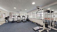 Located at 4637 Cason Cove Dr in Orlando, Florida, this single-story fitness and recreation facility offers a well-rounded environment for health and wellness activities. The space is organized around a grand central corridor finished with polished travertine tile flooring and soaring vaulted ceilings supported by white columns. This bright hallway connects the key areas of the facility and features glass-paneled doors that provide visual access to interior spaces, including a vibrant racquetball court with colorful wall panels visible through the glazing.

The main gym area is generously proportioned, carpeted in durable blue flooring, and lined with large multi-pane windows on multiple walls that allow abundant natural light and views of the surrounding greenery. The workout floor is equipped with a comprehensive selection of strength-training machines, free weights, benches, and a dedicated cardio section featuring treadmills, an elliptical, and a stationary bike. A full-length mirror along one wall amplifies the sense of space and supports form-checking during exercise. Recessed lighting throughout supplements the natural light for a well-illuminated environment. The facility presents a clean, functional, and welcoming atmosphere suited to a wide range of fitness needs.