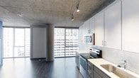 This contemporary single-floor space showcases an industrial-modern aesthetic defined by its striking exposed concrete ceiling, which adds raw architectural character throughout. Track lighting runs along the ceiling, providing flexible, directed illumination across the open layout. A bold structural concrete column anchors the living area, reinforcing the space's industrial design language.

The kitchen is thoughtfully appointed with crisp white upper cabinetry, dark lower cabinets, and light countertops that create a clean, contrasting palette. Subway tile backsplash adds a classic yet modern touch, while stainless steel appliances — including a range with oven and a built-in microwave — deliver a fully functional cooking environment. Dark hardwood flooring runs throughout, grounding the space with warmth against the cooler concrete overhead.

The standout feature is the expansive floor-to-ceiling window wall that floods the interior with abundant natural light and frames sweeping views of the surrounding urban skyline. The open-plan design seamlessly connects the kitchen and living areas, enhancing the sense of spaciousness. A built-in storage unit is also present along one wall, adding practical functionality. Overall, the space exudes a sophisticated urban atmosphere with a refined industrial edge.