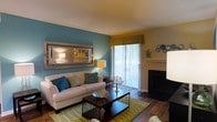 This single-story space showcases a thoughtfully designed living room that combines modern style with a warm, inviting ambiance. The room's most striking feature is a bold teal accent wall, which serves as a vibrant backdrop and adds depth and personality to the space. A large sliding glass door allows abundant natural light to pour in, illuminating the rich hardwood flooring that runs throughout the room.

The living area is generously proportioned, providing ample space for comfortable seating and furnishings. A plush white sofa, accented with teal and green throw pillows, anchors the seating arrangement, complemented by a dark wood coffee table set atop a patterned area rug in neutral and green tones. A large framed mirror is mounted on the teal wall, enhancing the sense of space and reflecting light throughout the room.

Vertical blinds dress the sliding glass door, offering privacy while still allowing light to filter in. Additional décor elements, including a floor lamp, wall-mounted decorative accents, and tasteful accessories, contribute to the cohesive and polished aesthetic of this well-appointed living space.