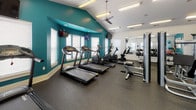 Located at 327 Guinevere Ln in Wilmington, NC, this fitness center is a well-appointed, single-floor facility designed to support a comprehensive workout experience. The space is characterized by a bold teal accent color on select walls, contrasting cleanly with the white walls and ceiling, creating an energizing and modern atmosphere throughout.

The cardio section is generously equipped with multiple treadmills from the 6000 Series Green line, as well as elliptical machines and stationary bikes, all arranged to allow comfortable spacing between users. Wall-mounted flat-screen TVs are positioned throughout the room to keep members entertained during workouts.

The strength training area offers a robust selection of equipment, including cable and pulley machines, multi-station cable systems, weight benches, a dumbbell rack with an accompanying mirror wall that amplifies the sense of space, and kettlebells. Resistance training accessories such as TRX suspension straps and stability balls further round out the available options.

The flooring throughout is a durable rubber gym surface, ideal for both heavy equipment and high-impact activity. Natural light filters in through multiple windows fitted with white blinds, while ceiling fans and bright overhead fluorescent lighting ensure the space remains well-ventilated and evenly lit. The vaulted ceiling in one section adds an open, airy quality to the overall environment.