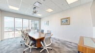 Located at 920 Manatee Avenue West in Bradenton, Florida, this professional conference room offers a well-appointed setting for meetings and collaborative work. The space is anchored by a substantial rectangular wooden conference table with a warm natural finish, surrounded by ergonomic mesh-back chairs in a contemporary white and blue-gray palette, accommodating up to ten attendees comfortably.

A large wall-mounted flat-screen display serves as a focal point for presentations, complemented by a wooden credenza beneath it for additional surface and storage. The room features a drop ceiling with recessed LED panels and a stylish circular pendant light fixture, ensuring consistent, professional illumination throughout.

Neutral gray carpeting and crisp white walls create a clean, distraction-free environment. Framed coastal artwork adds a touch of local character to the interior. The standout feature of this conference room is its floor-to-ceiling sliding glass windows, which frame sweeping panoramic views of the Bradenton waterfront and surrounding cityscape, infusing the workspace with abundant natural light and an inspiring backdrop. The combination of modern furnishings, presentation technology, and exceptional views makes this a highly functional and visually appealing meeting space.