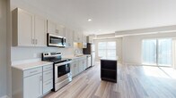This single-floor residence showcases a thoughtfully designed open-concept layout that seamlessly connects the kitchen and living areas. The kitchen is a standout feature, equipped with stainless steel appliances including a range, over-the-range microwave, dishwasher, and refrigerator. Light gray shaker-style cabinetry with brushed nickel hardware pairs elegantly with crisp white countertops. A contrasting dark-toned island with open shelving provides additional prep space and storage. Light wood-look luxury vinyl plank flooring flows throughout the main living areas, adding warmth and continuity. The living area is generously proportioned, bathed in natural light through sliding glass doors and multiple windows that invite outdoor views. The bedroom is carpeted for comfort and features a spacious walk-in closet with wire shelving and a window for added brightness. Two bathrooms serve the unit: one offers a full soaking tub and shower combo, while the other features a dedicated walk-in shower. Both bathrooms are finished with gray vanities, white countertops, and matching wood-look flooring. The overall aesthetic is clean and contemporary, with a neutral color palette of soft grays and warm whites creating a bright, welcoming atmosphere throughout.
