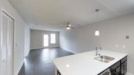 This single-story space showcases a clean, modern design with an open-concept layout that seamlessly connects the kitchen and living area. The kitchen is outfitted with dark cabinetry contrasted by a bright white countertop, a stainless steel sink with a contemporary gooseneck faucet, and a stainless steel dishwasher. A pendant light hangs above the kitchen island, providing focused task lighting while adding a stylish touch to the space. Sliding closet doors with a classic panel design are positioned along one wall near the kitchen, offering practical storage. The living area extends generously from the kitchen, featuring dark hardwood-style flooring that runs throughout and neutral gray walls that create a calm, cohesive atmosphere. A ceiling fan with a light fixture is centrally positioned in the living area, enhancing air circulation and ambient lighting. A set of double doors at the far end of the living space allows natural light to filter in and provides access to the outdoors. The textured ceiling adds subtle architectural character, and the overall design strikes a balance between functionality and contemporary style.