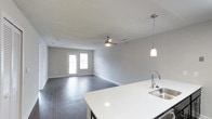 This single-story space showcases a clean, modern design with an open-concept layout that seamlessly connects the kitchen and living area. The kitchen is outfitted with dark cabinetry contrasted by a bright white countertop, a stainless steel sink with a contemporary gooseneck faucet, and a stainless steel dishwasher. A pendant light hangs above the kitchen island, providing focused task lighting while adding a stylish touch to the space. Sliding closet doors with a classic panel design are positioned along one wall near the kitchen, offering practical storage. The living area extends generously from the kitchen, featuring dark hardwood-style flooring that runs throughout and neutral gray walls that create a calm, cohesive atmosphere. A ceiling fan with a light fixture is centrally positioned in the living area, enhancing air circulation and ambient lighting. A set of double doors at the far end of the living space allows natural light to filter in and provides access to the outdoors. The textured ceiling adds subtle architectural character, and the overall design strikes a balance between functionality and contemporary style.