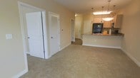 This single-floor residence offers a thoughtfully designed open-concept layout that seamlessly connects the main living area to the kitchen. The living space features neutral-toned carpeting that adds warmth and comfort underfoot, while the soft beige walls create a bright, welcoming ambiance throughout.

The kitchen is a functional highlight of the unit, equipped with light wood cabinetry, a black refrigerator, and a raised breakfast bar with a stone-style countertop — ideal for casual dining or entertaining. Elegant pendant lights hang above the bar, providing both task lighting and a refined aesthetic touch.

A series of interior doors along the hallway lead to additional rooms, suggesting a well-organized floor plan with clear separation between living and private spaces. A louvered closet door near the living area offers convenient storage access, while a paneled door at the end of the corridor provides passage to further rooms within the unit.

The consistent use of white trim and paneled doors throughout lends the space a clean, cohesive look. The neutral color palette and open flow make this residence a versatile canvas, easily adaptable to a variety of interior styles and furnishing preferences. Overall, the space balances practicality with an inviting, comfortable atmosphere.