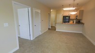 This single-floor residence offers a thoughtfully designed open-concept layout that seamlessly connects the main living area to the kitchen. The living space features neutral-toned carpeting that adds warmth and comfort underfoot, while the soft beige walls create a bright, welcoming ambiance throughout.

The kitchen is a functional highlight of the unit, equipped with light wood cabinetry, a black refrigerator, and a raised breakfast bar with a stone-style countertop — ideal for casual dining or entertaining. Elegant pendant lights hang above the bar, providing both task lighting and a refined aesthetic touch.

A series of interior doors along the hallway lead to additional rooms, suggesting a well-organized floor plan with clear separation between living and private spaces. A louvered closet door near the living area offers convenient storage access, while a paneled door at the end of the corridor provides passage to further rooms within the unit.

The consistent use of white trim and paneled doors throughout lends the space a clean, cohesive look. The neutral color palette and open flow make this residence a versatile canvas, easily adaptable to a variety of interior styles and furnishing preferences. Overall, the space balances practicality with an inviting, comfortable atmosphere.