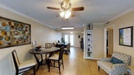 This single-floor space offers an open, flowing layout that seamlessly connects the dining and living areas. The interior is finished with warm hardwood-style flooring that runs throughout, complementing the neutral beige walls and crisp white crown molding that frames the ceiling. A ceiling fan with an integrated light fixture anchors the main living area, providing both functional lighting and air circulation.

The dining area accommodates a dark-toned round table with matching chairs, offering a cozy yet practical setting. Beyond the dining space, a secondary seating or workspace area is visible, benefiting from natural light that pours in through a set of glass French doors at the rear of the space. These doors provide an inviting connection to the outdoors while brightening the entire interior.

A built-in white shelving unit stands against one wall, offering decorative display space and adding a structured architectural element to the room. Crown molding detail further enhances the unit's integration into the overall design. The space maintains a clean, well-organized ambiance, making it functional and comfortable throughout.