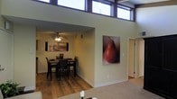 This single-floor space makes a striking architectural statement with its dramatically tall ceilings and a row of clerestory windows positioned near the roofline, flooding the interior with abundant natural light while maintaining privacy. The open layout creates a seamless visual connection between the living and dining areas, enhancing the sense of spaciousness.

The dining area is a well-defined zone, partially framed by a structural wall that adds subtle separation without fully enclosing the space. It features warm hardwood flooring, a ceiling fan with integrated lighting, and comfortably accommodates a dark wood dining table surrounded by matching chairs. Wall art adds a personal touch to the area.

Transitioning into the adjoining living space, the flooring shifts to carpet, providing a softer, more relaxed atmosphere. A large-scale canvas artwork of a pear serves as a bold focal point on the accent wall, contributing warmth and character to the neutral color palette of beige and cream tones that runs throughout.

A hallway visible near the accent wall suggests additional rooms or functional spaces beyond. Overall, the space balances modern architectural elements with warm, inviting finishes.