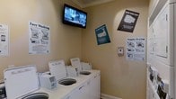 This communal laundry facility is designed with functionality and user convenience in mind. The single-floor space features a clean, welcoming layout with warm neutral-toned walls and classic crown molding details that lend a polished finish to the utilitarian setting.

The room is outfitted with a row of commercial-grade top-load washing machines, providing ample capacity for residents' laundry needs. Informational posters are displayed prominently on the walls, offering helpful guidance on operating the washers and dryers, tackling stains, and getting the most out of each cycle — making the space accessible and user-friendly for all.

A wall-mounted flat-screen television is positioned above the machines, allowing guests to stay entertained while waiting for their laundry. A payment or control panel is mounted on the side wall, ensuring a streamlined, self-service experience.

The overall ambiance is bright, tidy, and efficiently organized, reflecting a commitment to providing a practical yet comfortable shared amenity. The space is well-maintained, with clearly labeled equipment and instructional materials that make it easy for users to navigate the facility with confidence.