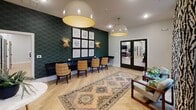 This thoughtfully designed single-floor space exudes a refined yet welcoming ambiance, blending contemporary style with warm, eclectic touches throughout. Light herringbone hardwood flooring anchors the main area, complemented by a series of large, statement pendant lights with gold interiors that cast a warm glow across the open layout. A striking deep teal patterned accent wall serves as a dramatic focal point, adorned with a curated gallery of framed artwork and flanked by brass wall sconces. A long built-in desk and upholstered chairs in graphic patterns line this wall, creating a functional yet stylish workspace. Sliding barn-style glass doors offer a sophisticated transition between spaces, while bold French doors with dark frames lead to adjacent rooms. A central seating arrangement features a marble-topped bistro table paired with woven rattan chairs, anchored by a vintage-style area rug that adds warmth and texture. A separate conference or meeting room boasts soaring ceilings, floor-to-ceiling windows with draped curtains, a dark wood table surrounded by woven chairs, and a glamorous gold multi-tiered chandelier overhead. Natural light filters in generously from multiple windows, offering views of the surrounding landscape and contributing to the inviting, upscale atmosphere of the space.