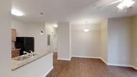This single-floor residence offers a thoughtfully designed open-concept layout that brings together the kitchen and living areas in a seamless, functional flow. The kitchen features a granite-style countertop peninsula with an integrated sink, a black refrigerator, and stylish pendant lighting — including a distinctive stained-glass accent pendant — that adds character to the space. Warm hardwood-style flooring runs throughout, grounding the interior with a cohesive, inviting feel. The living area is bathed in soft ambient light from an elegant bowl pendant fixture, creating a welcoming atmosphere. Neutral, light-toned walls amplify the sense of openness and allow for versatile interior styling. A hallway visible from the main living space leads to additional rooms, hinting at a practical and well-organized floor plan. An electrical panel is discreetly tucked along the hallway wall, and a white interior door provides access to further areas of the unit. Clean white baseboards frame the perimeter, adding a crisp finishing detail throughout. Overall, the space strikes a balance between modern practicality and comfortable residential living.