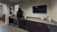 Located at 430 Allied Place in Gaithersburg, Maryland, this single-floor amenity space is thoughtfully designed to serve as a functional and welcoming community hub. The layout seamlessly blends a business center with casual lounge areas, creating a versatile environment suited for both focused work and relaxed socializing.

The business center area features a built-in, L-shaped granite-topped desk with dark cabinetry, providing ample workspace for multiple users. Computer workstations are set against neutral-toned walls adorned with contemporary abstract artwork, while large windows fitted with horizontal blinds and patterned drape panels allow natural light to fill the room.

Adjoining the workspace is a comfortable lounge area furnished with upholstered club chairs in warm gray and taupe tones, accented with nailhead trim detailing. A cylindrical metallic side table and a high-top bistro table with bar stools add to the relaxed, modern aesthetic. Framed mirrors on the walls enhance the sense of openness throughout the space.

The kitchenette area is equipped with granite countertops, dark lower cabinetry, a commercial-grade coffee machine, a stainless steel under-counter refrigerator, and a wall-mounted flat-screen television — ideal for casual gatherings. Textured carpeting throughout the main areas and tile flooring in the kitchenette complete the polished, contemporary design.
