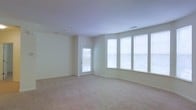 This single-floor space offers a clean, contemporary layout designed to maximize comfort and natural light. The main living area is a standout feature, highlighted by a sweeping bank of four large windows fitted with blinds that bathe the room in an abundance of diffused daylight. A glass-paneled door provides additional light and access to the exterior, reinforcing the bright and airy ambiance throughout.

The room is finished with soft, neutral-toned wall-to-wall carpeting that adds warmth underfoot and complements the light, creamy wall color. The smooth, flat ceiling is equipped with recessed ventilation and lighting fixtures, contributing to a clean and uncluttered aesthetic. The open floor plan provides generous, flexible living space that can be configured to suit a variety of needs.

The overall design language is understated and functional, favoring neutral tones and simple finishes that create a versatile backdrop for any interior style. The combination of ample natural light, open layout, and soft carpeting gives this space a welcoming and tranquil atmosphere well-suited for everyday living.