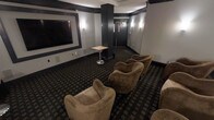 This private screening room offers a refined, theater-inspired environment designed for an immersive entertainment experience. The space is anchored by a large, framed wall-mounted display on one end, providing a commanding focal point for viewers. Deep charcoal and dark-toned walls, accented by warm wall sconces, create a dramatic and intimate atmosphere reminiscent of an upscale cinema lounge. The flooring features a patterned carpet in dark tones that complements the room's sophisticated color palette. Generous, curved lounge-style armchairs in warm tan upholstery are arranged in a tiered, theater-style layout, offering comfortable sightlines to the screen from every seat. A sleek pedestal side table provides a convenient surface for guests. Structural columns with dark cladding add an architectural dimension to the space while subtly reinforcing the room's design theme. At the far end, double doors provide entry and exit access, flanked by wall-mounted lighting that extends the room's warm, ambient glow into the entryway corridor. Overall, the space balances functionality with a polished, lounge-like aesthetic, making it well-suited for private screenings, presentations, or group entertainment gatherings.
