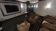 This private screening room offers a refined, theater-inspired environment designed for an immersive entertainment experience. The space is anchored by a large, framed wall-mounted display on one end, providing a commanding focal point for viewers. Deep charcoal and dark-toned walls, accented by warm wall sconces, create a dramatic and intimate atmosphere reminiscent of an upscale cinema lounge. The flooring features a patterned carpet in dark tones that complements the room's sophisticated color palette. Generous, curved lounge-style armchairs in warm tan upholstery are arranged in a tiered, theater-style layout, offering comfortable sightlines to the screen from every seat. A sleek pedestal side table provides a convenient surface for guests. Structural columns with dark cladding add an architectural dimension to the space while subtly reinforcing the room's design theme. At the far end, double doors provide entry and exit access, flanked by wall-mounted lighting that extends the room's warm, ambient glow into the entryway corridor. Overall, the space balances functionality with a polished, lounge-like aesthetic, making it well-suited for private screenings, presentations, or group entertainment gatherings.