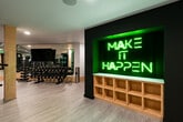 This contemporary fitness centre offers a comprehensive workout environment designed to accommodate a wide range of training styles. The space is accessed through sleek black-framed glass doors, which open into a bright, open-plan floor finished with light wood-effect flooring throughout. Natural wood-clad structural columns are evenly distributed across the floor, contributing to a clean, modern aesthetic while defining distinct workout zones. Recessed ceiling lighting ensures the entire space is well-lit, complemented by exposed industrial-style ductwork that adds an urban character. One wall features a bold botanical-inspired mural in green and white tones, serving as a striking visual focal point and reinforcing the energetic ambiance. Floor-to-ceiling mirrors line key sections of the facility, visually expanding the space and supporting form-checking during workouts. The cardio zone is equipped with multiple treadmills, rowing machines, and stationary bikes, while the strength area provides a full rack of dumbbells, weight benches, and multi-functional cable and power rack systems. A dedicated functional training station includes battle ropes and a punch bag. The facility includes clearly posted occupancy guidelines and digital access controls at the entrance, reflecting a well-managed and professional fitness environment.