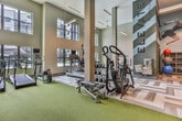 Located at 900 Battery Ave SE in Atlanta, Georgia, this fitness center offers a thoroughly equipped, single-floor workout facility designed for a comprehensive exercise experience. The space is defined by its soaring double-height ceilings, which create an open, airy atmosphere that is further enhanced by large, multi-pane windows along the perimeter walls. These windows flood the interior with natural light and provide views of the outdoor surroundings, including a glimpse of a pool area.

The flooring features a stylish chevron-patterned luxury vinyl tile, transitioning to green turf in the dedicated strength and functional training zone. Full-length mirrors along one wall visually expand the space and support form-checking during workouts. Equipment is thoughtfully arranged and includes Matrix elliptical cross-trainers, a StairMaster stepmill, treadmills, and a Life Fitness Smith machine with plate storage. A tiered kettlebell rack, a full dumbbell rack with bench, resistance training stations, and a wall-mounted target board round out the functional fitness offerings. Stability balls and additional accessories are neatly stored along the walls. Multiple wall-mounted flat-screen televisions are positioned throughout for entertainment. The overall design is modern and clean, with a neutral color palette accented by dynamic diagonal graphic details on the upper walls.