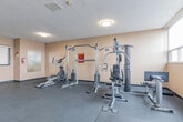 Located at 140 Robinson Street in Hamilton, Ontario, this fitness centre offers a well-organized, single-floor exercise space designed to meet a range of workout needs. The room features durable grey rubber tile flooring throughout, providing a safe and practical surface for all types of training activity.

Natural light streams in through a wide bank of horizontal windows along one wall, creating a bright and open atmosphere. The windows offer outward views, adding to the sense of space within the room. Overhead, flush-mount ceiling fixtures supplement the natural light, ensuring the space is well-lit at all times.

The fitness centre is equipped with a variety of exercise equipment thoughtfully arranged to maximize usable floor space. Cardio equipment includes multiple Spirit-brand treadmills and a stationary cycle, while strength training is supported by a multi-station cable machine and a flat utility bench. A freestanding dumbbell rack stocked with a range of weights provides options for free weight training. Wall-mounted mirrors are positioned near the entrance, allowing users to monitor their form during workouts.

The entrance features a glass-panelled door, and the warm beige-toned walls contribute to an inviting, functional ambiance. A fire extinguisher and safety signage are also present, reflecting attention to occupant safety. Overall, this fitness centre is a practical and well-appointed amenity space.