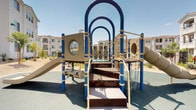 This residential community amenity space offers a thoughtfully designed outdoor environment centered around comfort, recreation, and community living. The grounds are landscaped with drought-tolerant plants, decorative gravel, and young trees that add greenery while requiring minimal upkeep — a practical and attractive choice for the sunny, arid climate.

A covered pavilion serves as a shaded gathering spot, featuring a ceiling fan, a round concrete table, and cylindrical concrete stools. The structure's white stucco columns and clean architectural lines complement the surrounding multi-story residential buildings, which share a consistent Mediterranean-inspired aesthetic with red tile rooflines, white exteriors, and black iron balcony railings.

Adjacent to the pavilion, a dedicated children's play area provides a vibrant and fully equipped playground structure with slides, climbing elements, interactive game panels, and chain accessories, all set on a cushioned rubberized surface for safety.

A designated BBQ station rounds out the amenities, featuring a freestanding gas grill on a concrete pad, positioned to take advantage of the open courtyard setting. A gated pool area is also visible in the background, further enhancing the community's recreational offerings. Together, these features create a functional and inviting outdoor space for residents of all ages.