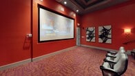 This dedicated screening room delivers an immersive cinematic experience through a thoughtfully designed, theater-style layout. Bold red walls set a dramatic tone throughout the space, complemented by a richly patterned carpet in deep crimson and gold hues. Rows of gray upholstered seats with integrated armrest cup holders are arranged in a tiered, stadium-style configuration, ensuring clear sightlines to the large projection screen mounted at the front of the room. A ceiling-mounted projector serves as the visual centerpiece, delivering crisp imagery to the generously sized framed screen. Acoustic treatment panels line one side wall, featuring a distinctive textured, cushioned design that aids in sound absorption and enhances audio clarity. Surround sound speakers are strategically positioned along the walls, complementing the room's acoustics. Warm wall sconces with ornate detailing provide ambient lighting between screenings, while recessed ceiling lights illuminate the space evenly. A central aisle with a handrail guides guests to upper rows via stepped tiers. Abstract black-and-white artwork adorns one wall, adding a refined artistic touch to the otherwise dramatic, cinema-inspired interior. The overall design balances functionality with a polished, upscale aesthetic.
