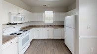 This single-story home offers a well-organized layout with three bedrooms and two full bathrooms, finished consistently throughout with wood-style flooring that lends warmth and cohesion to every room. Crown molding and ceiling fans with light fixtures are present across the living areas and bedrooms, contributing to a clean, classic aesthetic.

The living room is bright and open, featuring crown molding and a sliding glass door that opens to a balcony, providing natural light and outdoor views. The kitchen is outfitted with white cabinetry, dark countertops, a double sink, and a full suite of white appliances including a range, microwave, dishwasher, and refrigerator, all positioned around a functional U-shaped layout with a window above the sink.

The bedrooms are comfortably sized, each featuring neutral walls, natural light from windows with blinds, and ceiling fans. At least one bedroom includes an open closet with wire shelving for organized storage. The two bathrooms each feature a tub-shower combo with white tile surround, a single vanity with white cabinetry, and wood-style flooring. One bathroom features a darker granite-style countertop, while the second offers a lighter finish, providing subtle distinction between the two spaces.
