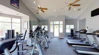 This fitness center offers a bright and open workout environment designed to support an active lifestyle. The space features soaring vaulted ceilings fitted with ceiling fans, which work alongside recessed lighting to ensure a well-ventilated and well-lit atmosphere throughout the day. Large windows and glass-paned doors line the walls, flooding the interior with abundant natural light and providing views of the surrounding greenery outside.

The workout floor is outfitted with a variety of cardio equipment, including elliptical trainers, treadmills, and additional aerobic machines, all arranged to allow comfortable movement and spacing between users. A full-length mirror along one wall enhances the sense of space while allowing members to monitor their form during exercise.

The interior palette is clean and neutral, with gray walls and striped carpeted flooring that lend a modern, professional feel to the space. The layout is straightforward and functional, with clear pathways to the exit and ample room to move between equipment. Overall, the fitness center strikes a balance between practicality and comfort, making it a welcoming space for a wide range of fitness routines.