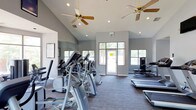 This fitness center offers a bright and open workout environment designed to support an active lifestyle. The space features soaring vaulted ceilings fitted with ceiling fans, which work alongside recessed lighting to ensure a well-ventilated and well-lit atmosphere throughout the day. Large windows and glass-paned doors line the walls, flooding the interior with abundant natural light and providing views of the surrounding greenery outside.

The workout floor is outfitted with a variety of cardio equipment, including elliptical trainers, treadmills, and additional aerobic machines, all arranged to allow comfortable movement and spacing between users. A full-length mirror along one wall enhances the sense of space while allowing members to monitor their form during exercise.

The interior palette is clean and neutral, with gray walls and striped carpeted flooring that lend a modern, professional feel to the space. The layout is straightforward and functional, with clear pathways to the exit and ample room to move between equipment. Overall, the fitness center strikes a balance between practicality and comfort, making it a welcoming space for a wide range of fitness routines.