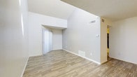 This single-floor space offers a clean and modern interior designed to maximize openness and comfort. The main living area immediately impresses with its vaulted ceilings, which rise dramatically to create a spacious, airy atmosphere that enhances the overall sense of volume. The soaring ceiling height draws the eye upward and contributes to an open, unencumbered feel throughout the space.

Underfoot, light wood-style flooring runs throughout the main area, lending warmth and a contemporary touch to the neutral palette. The walls are finished in a soft, light gray tone that complements the flooring and enhances the brightness of the interior. White baseboards provide a crisp contrast, contributing to a well-maintained and polished aesthetic.

The space is equipped with multiple HVAC vents, including both ceiling-mounted supply vents and floor-level return vents, ensuring efficient climate control throughout. A thermostat is conveniently positioned on the wall for easy access.

A vertical blind-covered door or window is set within a defined architectural recess, adding subtle structural character to the layout. The textured ceiling adds a classic finishing detail. Overall, this space presents a versatile, move-in-ready interior that balances practicality with an inviting, light-filled ambiance.