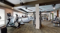 This modern fitness center is designed to support a full range of workout routines within a spacious, single-floor layout. The cardio zone is lined with large windows fitted with roller shades, allowing ample natural light while offering glare control. Rows of treadmills, elliptical trainers, and a stair climber are neatly arranged in this area, each machine equipped with an individual display screen. Industrial-style ceiling fans are mounted overhead to maintain airflow throughout the space.

The strength training section houses a comprehensive selection of cable machines and selectorized equipment, organized along mirrored walls that enhance the sense of depth and openness. Decorative elements, including natural wood slat partitions and hexagonal moss wall art, add a modern, biophilic aesthetic to the environment.

A dedicated functional training area provides plyometric boxes in multiple sizes, kettlebells, medicine balls, sandbags, and stability balls, all stored on wall-mounted racks for easy access. Rowing machines and a spin bike round out the cardio offerings in this zone. The flooring throughout consists of textured, multi-toned carpet tiles that are both durable and visually cohesive. Recessed and pendant lighting complement the overall design, creating a bright and motivating atmosphere suited for a wide variety of fitness activities.