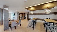 This single-floor amenity space is thoughtfully designed with a contemporary aesthetic, combining functionality and style across several distinct zones. Light wood-look flooring runs continuously throughout, unifying the open layout and contributing to the bright, airy atmosphere.

The lounge area serves as a central gathering point, furnished with plush wingback chairs, a sofa, and a wooden coffee table arranged around a wall-mounted flat-screen TV and a sleek linear fireplace set into a striking textured wave-panel accent wall. Large glass doors in this area provide direct access to an outdoor pool, flooding the space with natural light.

Adjacent to the lounge, a spacious dining and social area features high-top communal tables paired with industrial-style metal stools, beneath an eye-catching coffered ceiling detail with integrated LED lighting and elegant crystal pendant chandeliers. Additional casual seating and standard-height tables provide flexible options.

The kitchen is equipped with a large granite-topped island, stainless steel appliances including a range with hood, a wine cooler, and a decorative geometric tile backsplash. A sliding glass door opens to an outdoor area, enhancing natural light.

A glass-enclosed business center with workstations and a private corridor with wood-clad columns complete this well-appointed communal space.