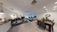 This single-floor community clubhouse is designed to offer a comfortable and functional gathering space with a clean, contemporary aesthetic throughout. The interior is unified by light-toned tile flooring, white beadboard wall paneling, and crown molding that lend a cohesive, polished character to every room.

The main lounge area is generously proportioned, featuring multiple seating arrangements anchored by a fireplace and a large flat-screen television. Bold, abstract area rugs add visual warmth against the neutral flooring, while recessed lighting ensures the space is well-illuminated. French doors at the rear of the lounge open directly to an outdoor patio and landscaped grounds, allowing abundant natural light to filter in and providing seamless indoor-outdoor connectivity.

A dedicated dining area sits adjacent to the lounge, furnished with a rich dark-wood table and upholstered chairs beneath recessed ceiling lights. A pair of large abstract canvas artworks adorns the white beadboard walls, contributing an artistic focal point to the space.

The kitchen is equipped with stainless steel appliances, white shaker-style cabinetry, dark granite countertops, and a pass-through bar counter illuminated by a trio of pendant lights—ideal for serving and social interaction. A built-in wine rack adds a refined detail to the cabinetry. Together, these spaces create a versatile and inviting environment suited for community gatherings and everyday use.