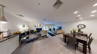 This single-floor community clubhouse is designed to offer a comfortable and functional gathering space with a clean, contemporary aesthetic throughout. The interior is unified by light-toned tile flooring, white beadboard wall paneling, and crown molding that lend a cohesive, polished character to every room.

The main lounge area is generously proportioned, featuring multiple seating arrangements anchored by a fireplace and a large flat-screen television. Bold, abstract area rugs add visual warmth against the neutral flooring, while recessed lighting ensures the space is well-illuminated. French doors at the rear of the lounge open directly to an outdoor patio and landscaped grounds, allowing abundant natural light to filter in and providing seamless indoor-outdoor connectivity.

A dedicated dining area sits adjacent to the lounge, furnished with a rich dark-wood table and upholstered chairs beneath recessed ceiling lights. A pair of large abstract canvas artworks adorns the white beadboard walls, contributing an artistic focal point to the space.

The kitchen is equipped with stainless steel appliances, white shaker-style cabinetry, dark granite countertops, and a pass-through bar counter illuminated by a trio of pendant lights—ideal for serving and social interaction. A built-in wine rack adds a refined detail to the cabinetry. Together, these spaces create a versatile and inviting environment suited for community gatherings and everyday use.