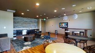 This single-floor space is a thoughtfully designed lounge and entertainment area that blends modern aesthetics with warm, inviting elements. The room is anchored by a striking reclaimed wood accent wall that adds texture and character to the space, complemented by rich hardwood flooring throughout. Recessed ceiling lighting provides ambient illumination, while a pendant light adds a decorative focal point overhead.

The lounge area is furnished with contemporary seating, including leather armchairs and accent chairs arranged around a low, round coffee table atop a patterned area rug, creating an intimate conversation zone. A wall-mounted flat-screen television serves as the entertainment centerpiece, positioned at eye level for comfortable viewing.

Along one wall, a built-in wet bar with dark cabinetry offers a functional hospitality station, ideal for entertaining. Adjacent high-top bar seating and a bistro-style table and chairs provide additional gathering options. The open, flowing layout allows for versatile use of the space, accommodating both casual relaxation and social gatherings. Neutral wall tones paired with the warm wood elements and modern furnishings create a sophisticated yet comfortable ambiance throughout.