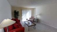 This single-floor space centers around a comfortable living room finished with plush, neutral-toned carpeting that adds warmth underfoot. A large sliding glass door with vertical blinds serves as the room's focal point, allowing generous natural light to fill the space and providing access to the outdoors. The walls are painted in a soft, neutral tone, contributing to an open and airy feel throughout.

The living room is furnished with a bold red sofa and a coordinating striped armchair with ottoman, creating a classic seating arrangement. An ornate wrought-iron and glass coffee table anchors the seating area, complemented by a matching side table topped with a traditional table lamp. A lush indoor plant in the corner adds a natural, refreshing element to the space.

The layout is practical and well-proportioned, with clean sightlines and an uncluttered design. The combination of natural light, neutral walls, and warm carpeting creates an inviting, relaxed atmosphere suited for everyday living and entertaining alike.
