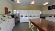 This shared laundry facility is a functional, well-organized space designed to accommodate multiple users efficiently. The room features ten coin-operated machines arranged in a row of stacked washer and dryer pairs along the main wall, numbered for easy identification. The machines are uniformly white and equipped with clearly labeled controls, making them straightforward to operate.

At the center of the room, a sturdy wooden folding table with a warm-toned surface provides ample space for sorting and folding laundry. Additional utility surfaces and wire laundry carts along the side walls offer further convenience for residents managing their loads.

The space is illuminated by flush-mounted ceiling light fixtures that cast even, practical lighting throughout the room. The walls are painted a neutral yellow tone, contributing to a clean and approachable atmosphere. Tile flooring in a neutral gray completes the utilitarian aesthetic while remaining easy to maintain.

Natural light supplements the artificial lighting through a window along one wall, enhancing the overall brightness of the space. The layout is thoughtfully arranged to allow multiple users to move comfortably between machines and the folding area simultaneously.