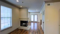 This two-story space combines classic architectural details with a warm, inviting atmosphere. The main living area immediately draws attention with its rich hardwood flooring, which adds depth and character throughout the open layout. A striking corner fireplace serves as the room's focal point, framed by a crisp white surround and raised hearth that lend an air of elegance and craftsmanship. Crown molding runs along the ceiling perimeter, adding a refined finishing touch that complements the neutral, light-toned walls. Recessed lighting is thoughtfully distributed across the ceiling, providing even, ambient illumination that enhances the brightness of the space. A set of French doors at the far end of the living area floods the room with natural light and offers a seamless visual connection to the outdoors. The open floor plan creates a generous sense of flow and space, making the area feel both comfortable and functional. Cabinetry is visible in an adjacent area, hinting at additional utility spaces nearby. Spanning two floors, this property offers versatile living arrangements with a cohesive design language carried throughout, defined by quality materials, thoughtful details, and a clean, timeless aesthetic.