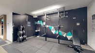 Located at 1100 Avenue du Docteur-Penfield in Montréal, this fitness facility offers a thoughtfully designed workout environment spanning a generous single-floor footprint. The main cardio and training area features a bright, open layout accentuated by exposed wooden ceiling beams and large windows that invite natural light, creating an airy and motivating atmosphere. The light-toned flooring complements the clean white walls, while full-length mirrors along one side enhance the sense of space. This section is outfitted with a row of True treadmills, elliptical trainers, stationary bikes, rowing machines, and a cable/multi-functional strength machine, catering to a wide range of fitness needs. A dedicated strength and free weights zone transitions seamlessly from the main area, distinguished by dark accent walls adorned with a striking geometric mural in teal, grey, and white. This space features rubber flooring for safety, a power rack with barbell and weight plates, an adjustable bench, resistance bands, and a water fountain. A glass-enclosed area provides additional storage or equipment access. Informational signage and workout guides are displayed throughout, making the facility user-friendly. The overall ambiance is modern, functional, and energizing, suitable for both casual exercisers and dedicated athletes.