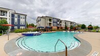 Located at 19221 Beechnut St in Richmond, Texas, this outdoor amenity space is centered around a generously sized, resort-style swimming pool with striking turquoise water. The pool features a freestanding spa with a bubbling water feature, in-pool lounge ledges for relaxing in the water, and a wide surrounding deck finished in neutral concrete. Blue chaise lounge chairs are arranged throughout the pool deck, providing ample space for sunbathing and leisure. Two blue canopy cabanas offer shaded retreats, complemented by red market umbrellas positioned at additional seating areas. The pool area is framed by manicured landscaping, including trimmed shrubs, ornamental grasses, and young trees that soften the hardscape and add greenery throughout. A pergola structure is visible along the perimeter, extending the outdoor gathering options. The adjacent clubhouse building presents a classic design with a combination of brick and siding exterior, pitched rooflines, and large windows that visually connect the interior to the pool deck. Multi-story residential buildings surround the community, providing a cohesive backdrop. The entire pool area is enclosed by a perimeter fence, ensuring a secure and well-defined recreational environment for residents.