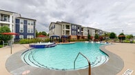 Located at 19221 Beechnut St in Richmond, Texas, this outdoor amenity space is centered around a generously sized, resort-style swimming pool with striking turquoise water. The pool features a freestanding spa with a bubbling water feature, in-pool lounge ledges for relaxing in the water, and a wide surrounding deck finished in neutral concrete. Blue chaise lounge chairs are arranged throughout the pool deck, providing ample space for sunbathing and leisure. Two blue canopy cabanas offer shaded retreats, complemented by red market umbrellas positioned at additional seating areas. The pool area is framed by manicured landscaping, including trimmed shrubs, ornamental grasses, and young trees that soften the hardscape and add greenery throughout. A pergola structure is visible along the perimeter, extending the outdoor gathering options. The adjacent clubhouse building presents a classic design with a combination of brick and siding exterior, pitched rooflines, and large windows that visually connect the interior to the pool deck. Multi-story residential buildings surround the community, providing a cohesive backdrop. The entire pool area is enclosed by a perimeter fence, ensuring a secure and well-defined recreational environment for residents.