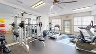 Located in Hickory, NC, this well-appointed fitness center offers a comprehensive exercise environment designed to support a wide range of workout routines. The single-floor facility features durable rubber flooring throughout, providing both safety and comfort during training sessions. Large windows along one wall allow natural light to fill the space, while a ceiling fan and fluorescent lighting ensure year-round comfort and visibility. The cardio section is equipped with multiple treadmills, elliptical trainers, a stationary recumbent bike, and a Precor cross-trainer, catering to users of varying fitness levels. For strength training, the facility includes a Smith machine with weight plates, a multi-function cable machine, a dedicated bench press setup, and a full rack of dumbbells. Floor-to-ceiling mirrors on one wall expand the sense of space and allow users to monitor their form during exercises. A wall-mounted rack holds a colorful assortment of medicine balls, resistance bands, and stability balls, rounding out the functional fitness offerings. A water fountain is conveniently accessible within the room. The clean, white walls and uncluttered layout contribute to an organized and motivating atmosphere, making this fitness center a practical and well-resourced facility for daily use.