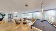 Located at 2001 Hollywood Boulevard in Hollywood, FL, this sophisticated single-floor living space offers a seamlessly flowing open layout with distinct functional zones designed for both relaxation and entertainment. Gleaming polished marble floors reflect abundant natural light throughout, lending the space an elegant and airy atmosphere.

The main lounge area features a curated mix of contemporary seating arrangements, including modular chairs and curved accent pieces, complemented by gold-toned side tables and wall-mounted sconces that add warmth to the bright white walls. A wall-mounted flat-screen television anchors one side of the room, while a striking black-and-white geometric accent wall defines a more intimate lounge nook furnished with a leather sectional sofa and a glass coffee table atop a textured area rug.

Floor-to-ceiling sheer curtain panels line an entire wall of windows, offering sweeping views of lush tropical palm trees and filling the space with soft, diffused light. A high-top bistro table with bar stools is positioned to take full advantage of this serene outdoor backdrop.

A billiards table provides a dedicated recreation area, while a sleek wet bar with dark stone countertops and white cabinetry serves as a functional centerpiece. The entry area is anchored by a geometric pendant light fixture, elegant navy accent chairs, and bold monochromatic wall art, creating a polished and welcoming first impression.