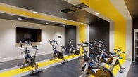 Located at 240 S Monaco Street Parkway in Denver, Colorado, this fitness and spin studio facility offers a purposefully designed layout across a single floor, encompassing a spin room, an open exercise studio, and a connecting hallway. The spin room is a standout feature, boldly decorated with striking yellow and dark gray striped flooring and matching ceiling accents that create a high-energy atmosphere. Multiple Stages brand stationary bikes are arranged in rows, complemented by recessed lighting and a wall-mounted monitor, making it well-suited for group cycling classes. The open exercise studio offers a clean, spacious environment finished with light wood-style flooring, bright recessed lighting, and a full wall of mirrors paired with a ballet barre — ideal for yoga, stretching, dance, or multi-purpose fitness classes. Along one wall, a well-organized equipment rack displays an array of dumbbells, medicine balls, resistance bands, foam rollers, and stability balls, set against a bold fitness-themed typographic mural that reinforces the space's active identity. The hallway connects both studios efficiently, providing a smooth flow throughout the facility. Together, the rooms deliver a comprehensive, professionally outfitted fitness environment designed to support a wide range of workout disciplines.