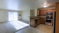 This single-floor residence offers a practical and comfortable open-concept layout that seamlessly connects the kitchen and living areas. The kitchen is well-appointed with warm honey-toned wood cabinetry, dark granite countertops, and a suite of stainless steel appliances including a dishwasher and range. A peninsula with a built-in sink serves as both a functional workspace and a natural divider between the kitchen and the adjoining living area. Tile flooring runs throughout the kitchen, providing a durable and easy-to-maintain surface. The living area transitions to plush wall-to-wall carpeting, adding warmth and comfort underfoot. Light neutral walls throughout create a clean, airy atmosphere that complements the space's open design. A large sliding glass door or wide window in the living area fills the space with abundant natural light, creating a bright and welcoming environment. A wall-mounted air conditioning unit ensures year-round comfort. The layout is efficient and cohesive, making it an ideal space for both everyday living and entertaining.