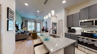 This single-story residence showcases a thoughtfully designed open-concept layout connecting the kitchen, dining, and living areas. The kitchen is a standout feature, equipped with gray shaker-style cabinetry, granite countertop surfaces, a white hexagonal tile backsplash, and a full suite of stainless steel appliances including a refrigerator with ice maker, range, and built-in microwave. A large central island with an undermount sink provides additional prep space and casual seating, illuminated by a cluster of pendant lights overhead. Light wood-style flooring flows throughout the main living areas, complementing the neutral color palette. The living room features a distinctive sage green accent wall, ceiling fan, and large windows with access to the outdoors, allowing generous natural light. Two bedrooms are finished with plush carpeting and bright windows; the primary bedroom includes its own accent wall and ample natural light. Each bathroom features granite-topped vanities with gray cabinetry, wood-style flooring, and direct access to a dedicated walk-in closet. One bathroom includes a glass-enclosed shower, while the primary bath offers a double vanity. Recessed lighting throughout enhances the clean, contemporary ambiance of the space.